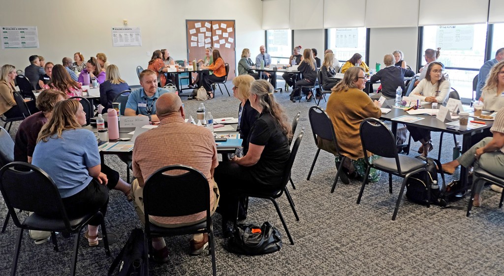 A busy conference room filled with people engaged in discussions around tables, collaborating on various topics.