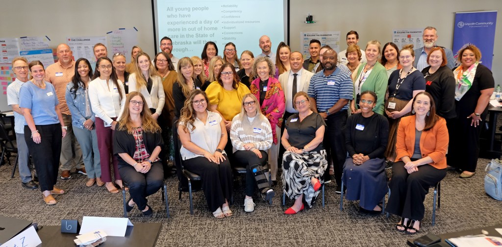 A large group photo of diverse individuals gathered for a meeting or event, smiling and standing together in a conference room with presentation materials in the background.