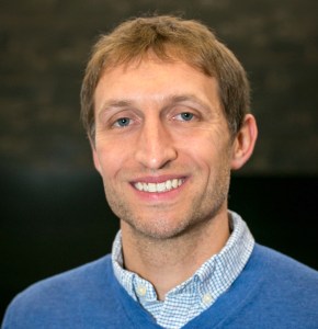 Smiling man with light brown hair, wearing a blue sweater and a collared shirt, against a blurred background.