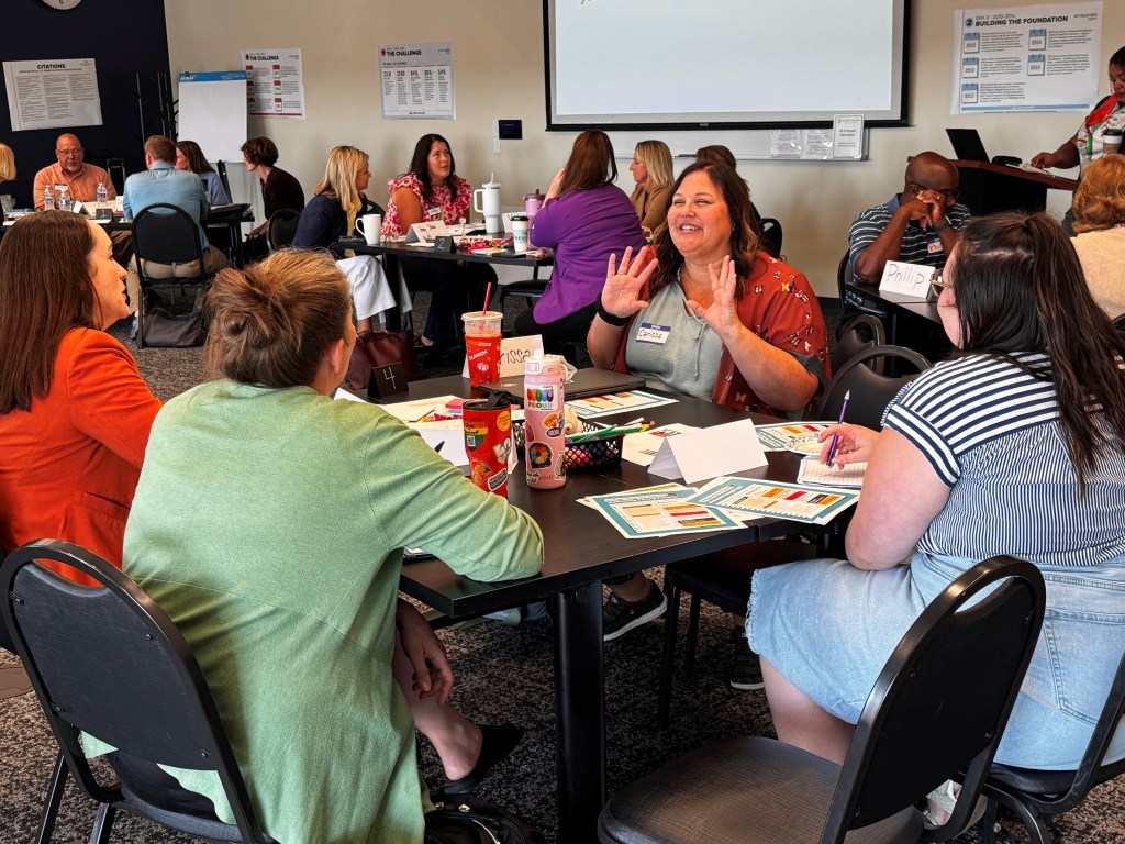 A group of people engaging in a collaborative discussion at tables in a meeting space, with a focus on a smiling woman in a floral shirt animatedly talking to her peers.