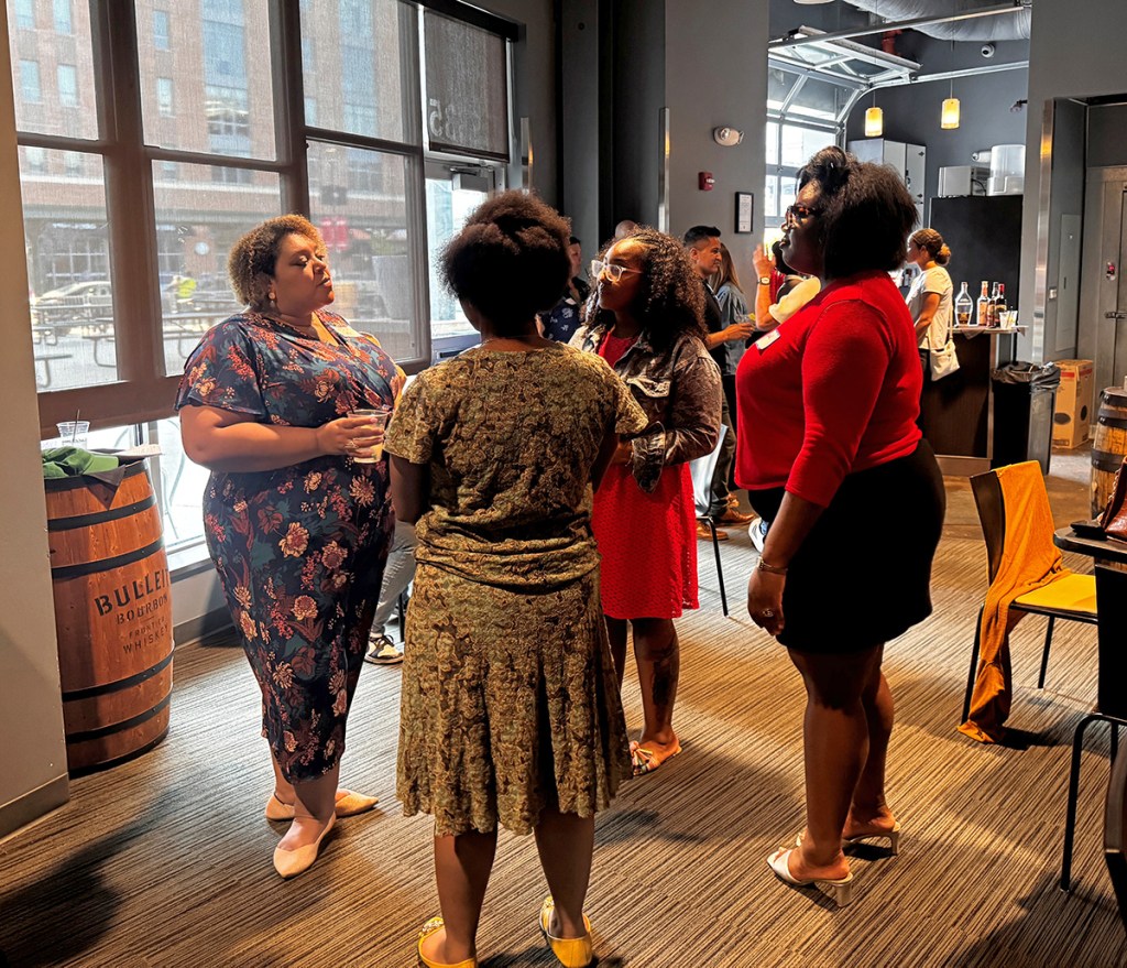 A group of five women engaging in conversation indoors, in a modern setting with large windows and wooden decor. One woman is holding a drink, while others are wearing colorful outfits. The atmosphere appears social and lively.