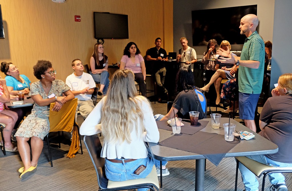 A group of people engaged in a discussion at a social event, with a speaker standing and addressing the audience in a casual setting.